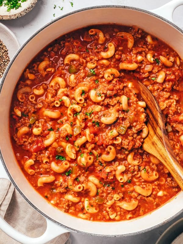 A white Dutch oven filled with American goulash made from tender elbow macaroni, crumbled ground beef, diced tomatoes, onions, and green bell peppers in a rich red sauce. Chopped fresh parsley is scattered over the pasta, and a wooden spoon rests inside the pot. Small bowls of parsley and black pepper sit nearby.