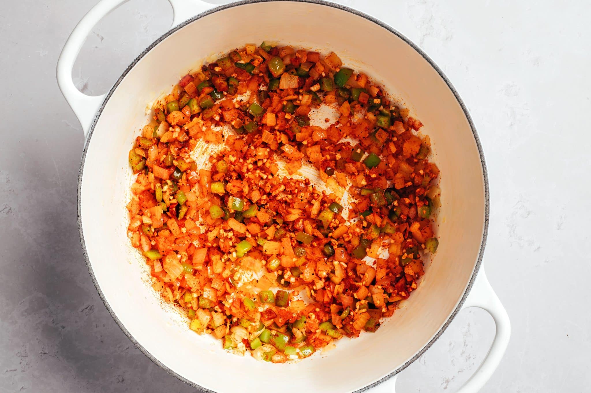 Softened diced onions and green bell peppers coated in reddish-orange paprika and black pepper inside a white Dutch oven. Small pieces of minced garlic are mixed throughout the vegetables, with a light coating of oil and seasonings across the bottom of the pot.