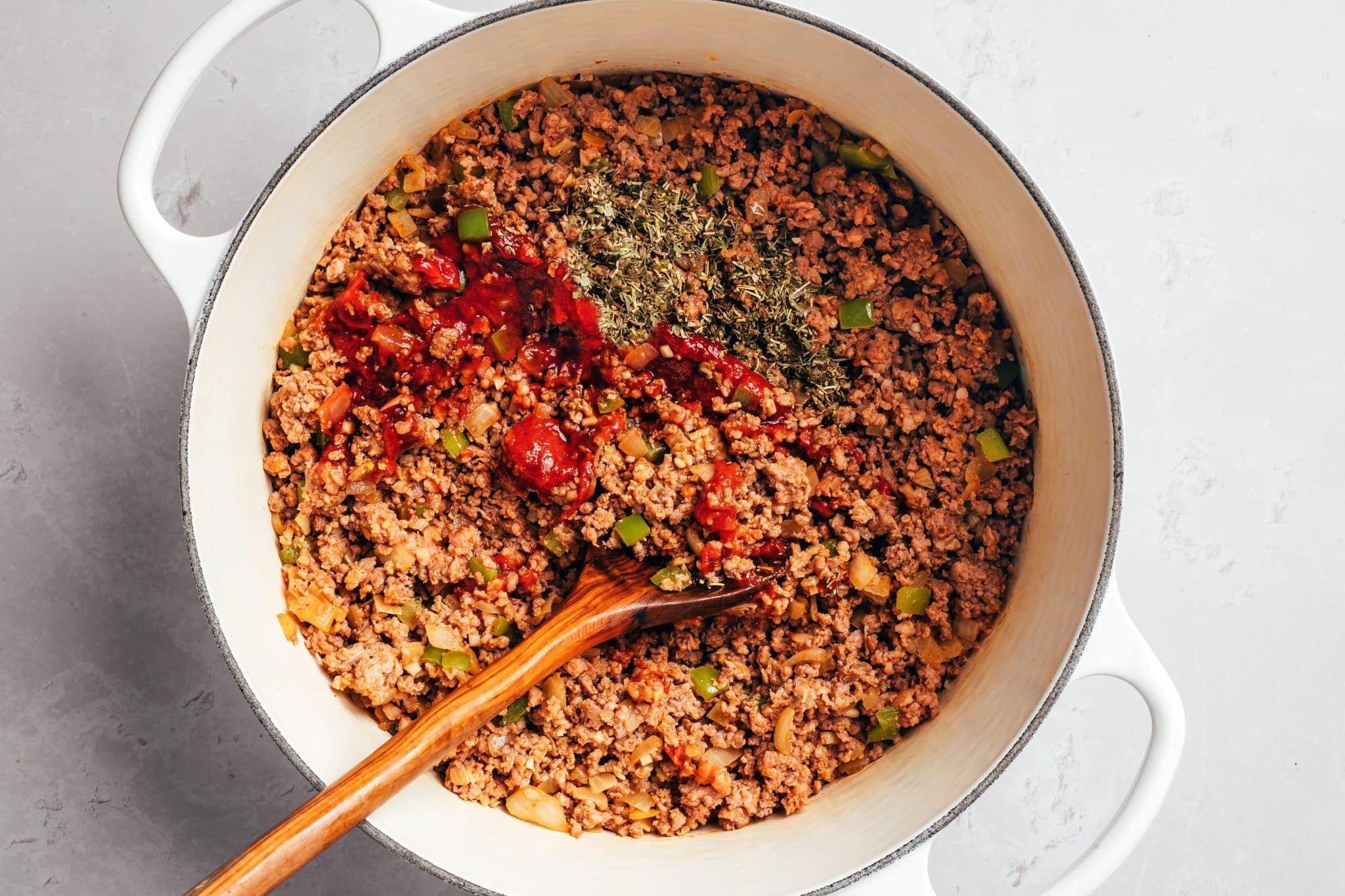 Browned ground beef mixed with diced onions and green bell peppers in a white Dutch oven. Thick red tomato paste and a mound of dried Italian seasoning sit on top of the beef, with a wooden spoon resting in the mixture.