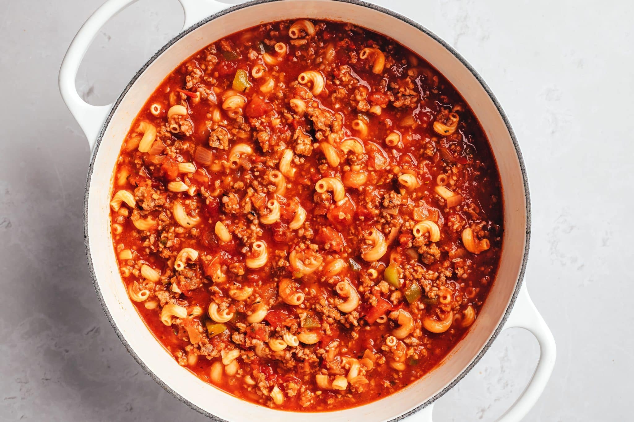 Curved elbow macaroni distributed throughout a thick, bright red tomato sauce with crumbled ground beef, diced tomatoes, onions, and small pieces of green bell pepper. The pasta is coated in sauce, with a small amount of liquid visible between the ingredients.