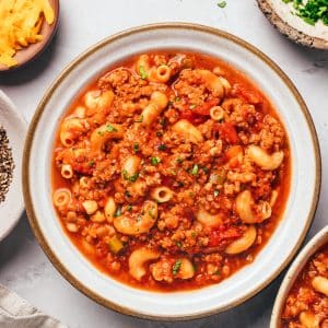 A shallow white ceramic bowl with a brown rim filled with American goulash. Tender elbow macaroni and crumbled ground beef are coated in a thick red tomato sauce with diced tomatoes, onions, and green bell peppers. Fresh chopped parsley is sprinkled over the pasta, with bowls of shredded cheddar and parsley nearby.