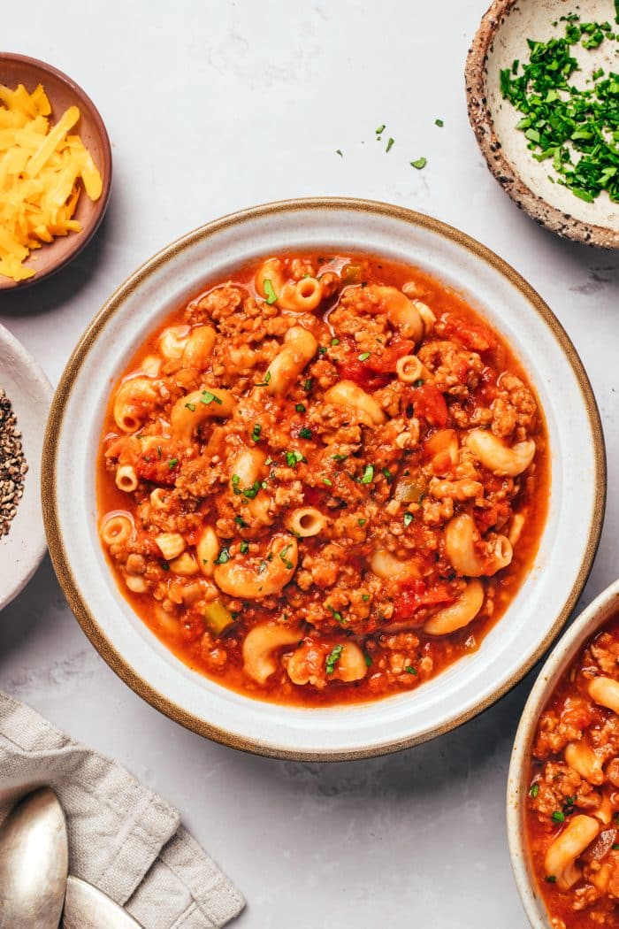 A shallow white ceramic bowl with a brown rim filled with American goulash. Tender elbow macaroni and crumbled ground beef are coated in a thick red tomato sauce with diced tomatoes, onions, and green bell peppers. Fresh chopped parsley is sprinkled over the pasta, with bowls of shredded cheddar and parsley nearby.