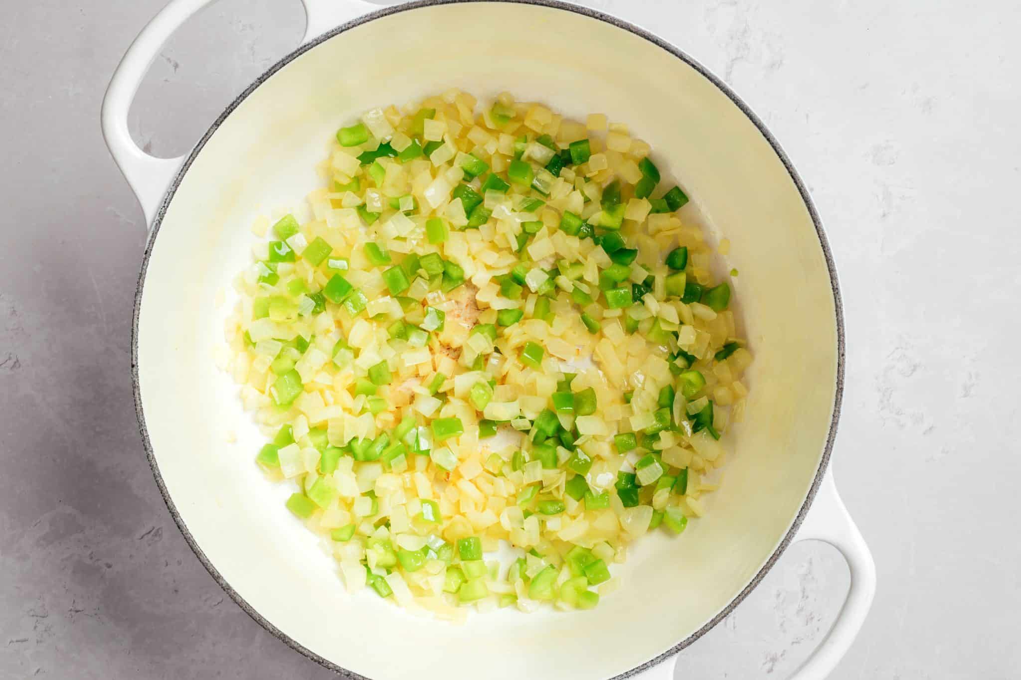 Diced yellow onions and green bell peppers softened in olive oil inside a white enameled Dutch oven. The onions are becoming translucent, while the peppers remain bright green and slightly tender.