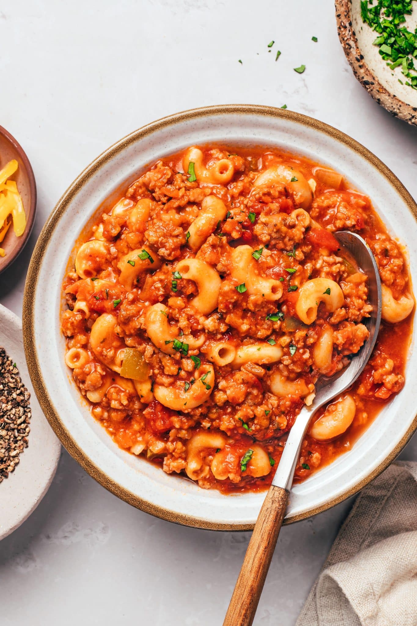 A white ceramic bowl filled with thick, saucy American goulash made with curved elbow macaroni, ground beef, diced tomatoes, and green bell peppers. A metal spoon with a wooden handle holds a generous portion of the beef and macaroni mixture along the right side of the bowl. Small pieces of fresh parsley are scattered across the pasta.