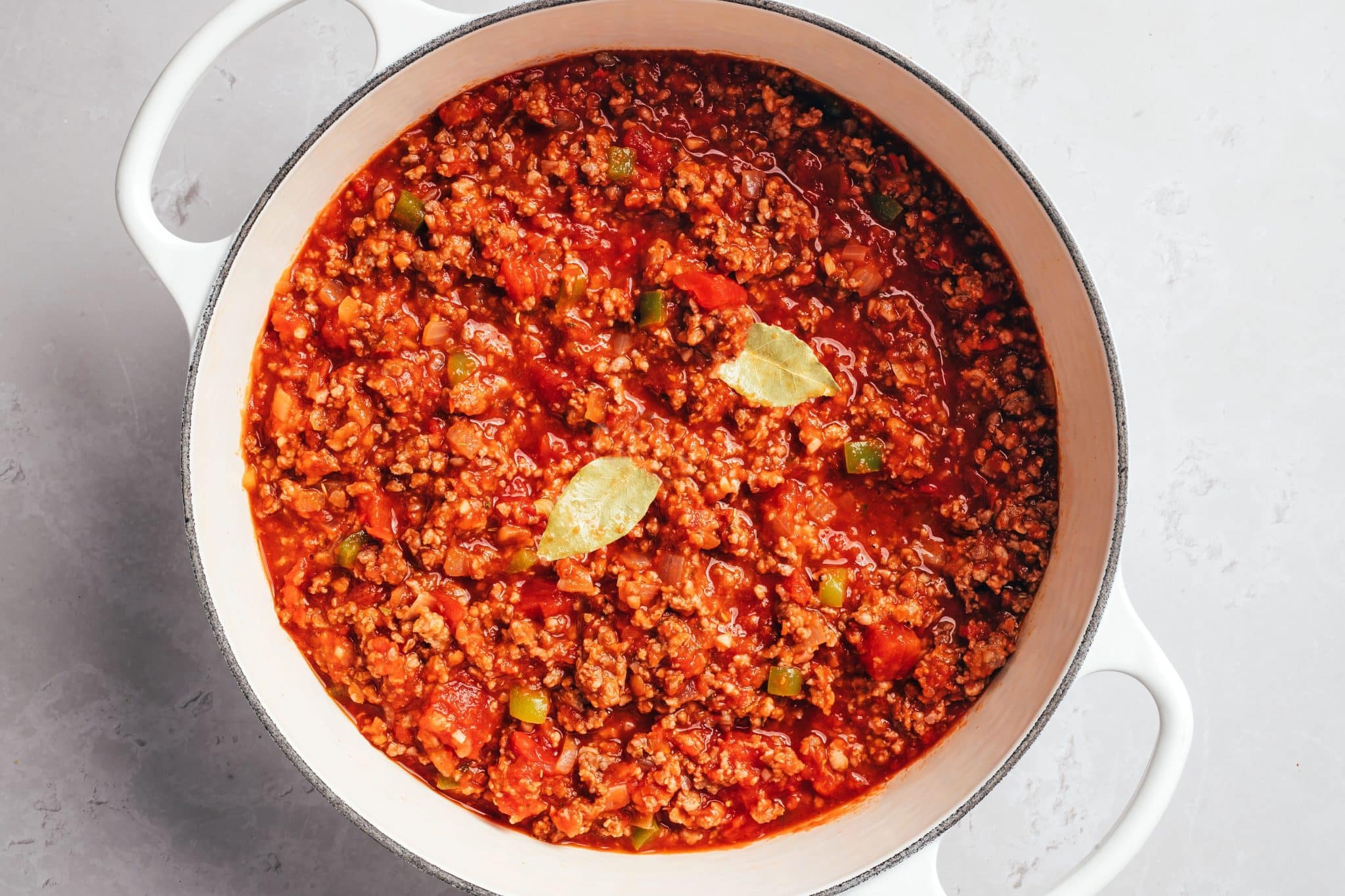 Thick red tomato sauce filled with crumbled ground beef, diced tomatoes, softened onions, and pieces of green bell pepper in a white Dutch oven. Two whole bay leaves rest on the surface of the chunky tomato-beef mixture.