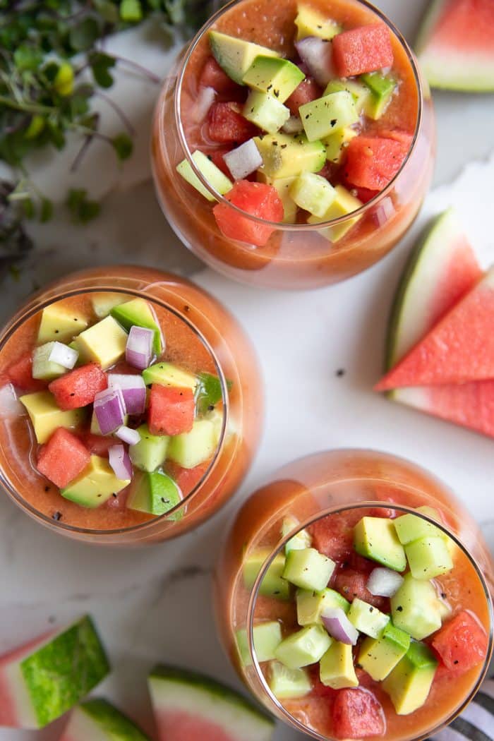 Overhead image of three glasses filled with watermelon gazpacho and topped with chopped avocado, cucumber, and red onion.