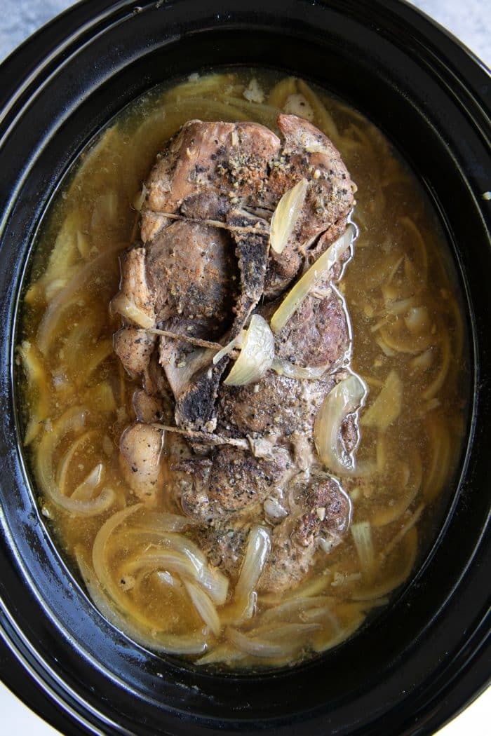 Fully cooked pork shoulder that is still wrapped in kitchen twine surrounded by a bath of cooked onions and apple cider.