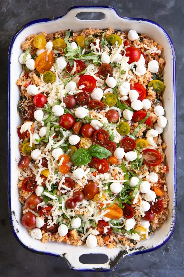 Uncooked casserole made with cauliflower rice, quinoa, chicken, kale, tomatoes, basil, and mozzarella cheese all topped with fresh cherry tomatoes, more mozzarella cheese, and fresh basil.