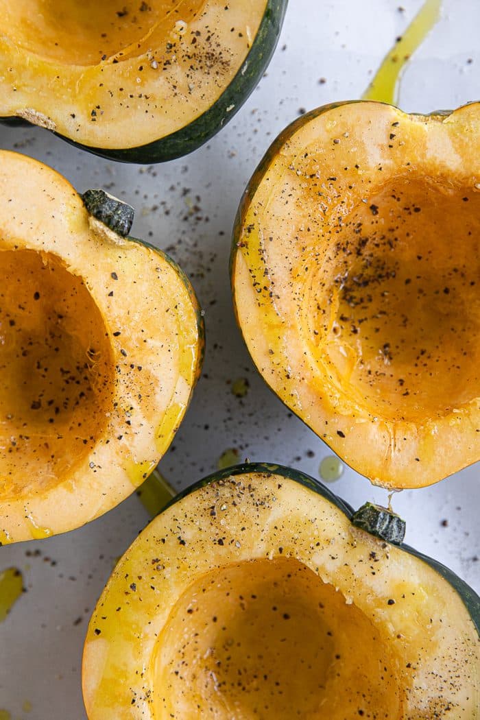 Raw halves of acorn squash on a baking sheet drizzled with olive oil and sprinkled with salt and pepper.