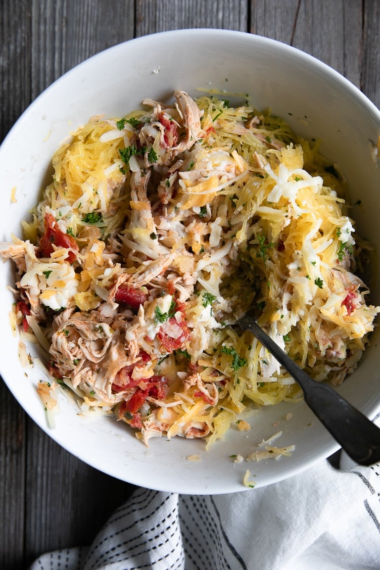 Large white bowl filled with mixed together shredded chicken and tomatoes, mixed shredded cheese, cooked spaghetti squash, chopped parsley, and ricotta cheese.