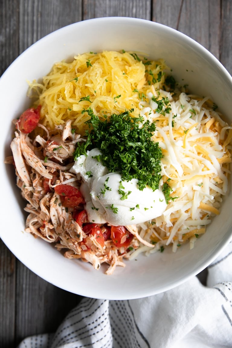 Large white bowl filled with shredded chicken and tomatoes, mixed shredded cheese, cooked spaghetti squash, chopped parsley, and ricotta cheese.