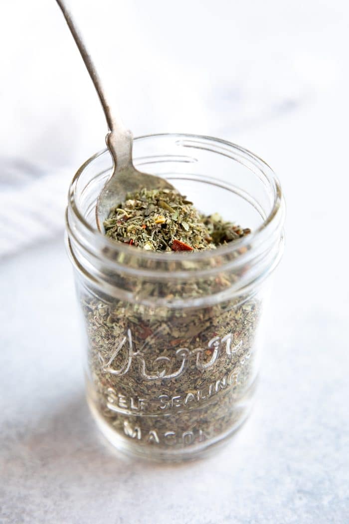 Spoon scooping a spoonful of dried Italian seasoning from a small mason jar.