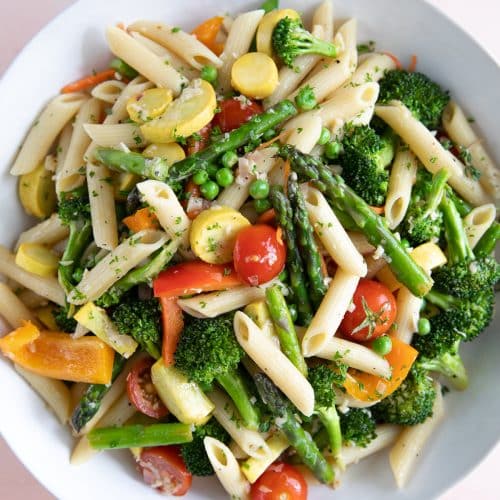 Large white pasta bowl filled with penne pasta and mixed spring vegetables such as asparagus, bell peppers, zucchini, tomatoes, and sprinkled with parmesan.