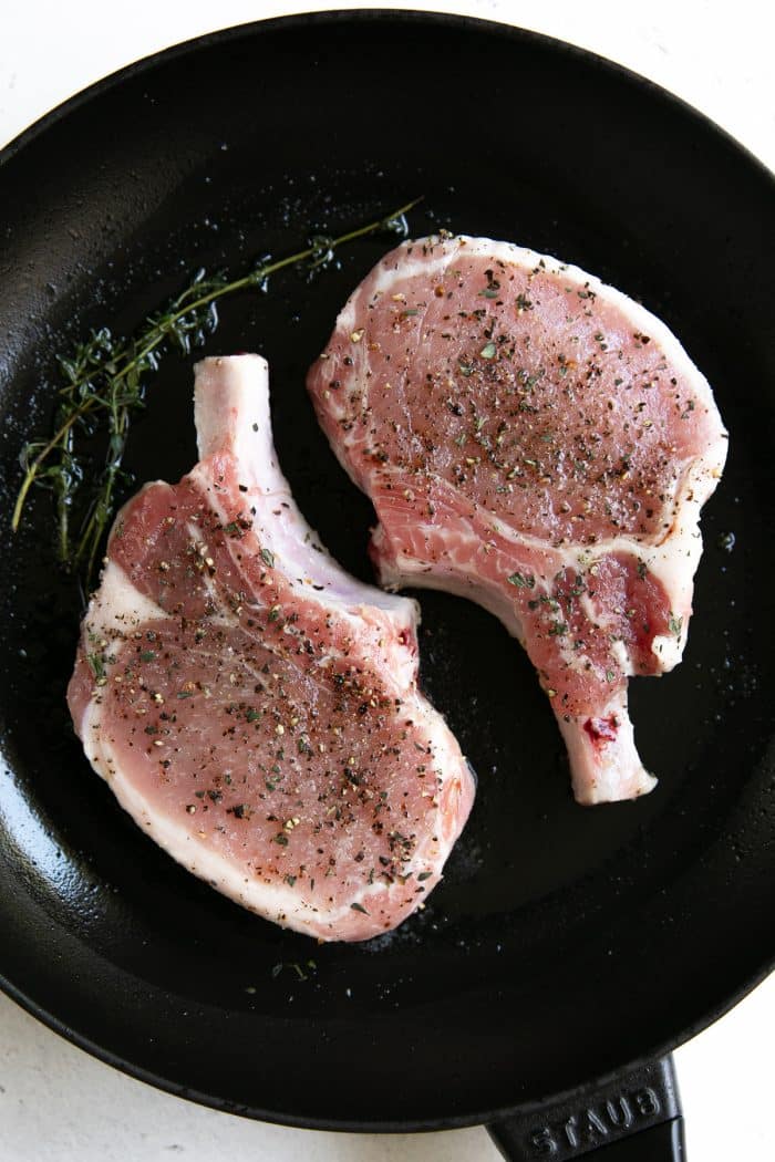 Two bone-in pork chops cooking in a medium cast iron skillet.