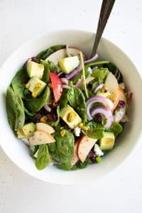 Salad bowl filled with baby spinach, sliced apples, avocado, sliced red onion, dried cranberries, pistachios, and feta cheese all tossed with a light red wine vinaigrette.
