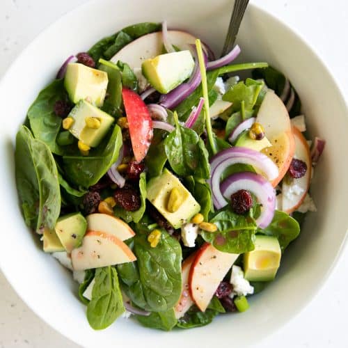 Salad bowl filled with baby spinach, sliced apples, avocado, sliced red onion, dried cranberries, pistachios, and feta cheese all tossed with a light red wine vinaigrette.
