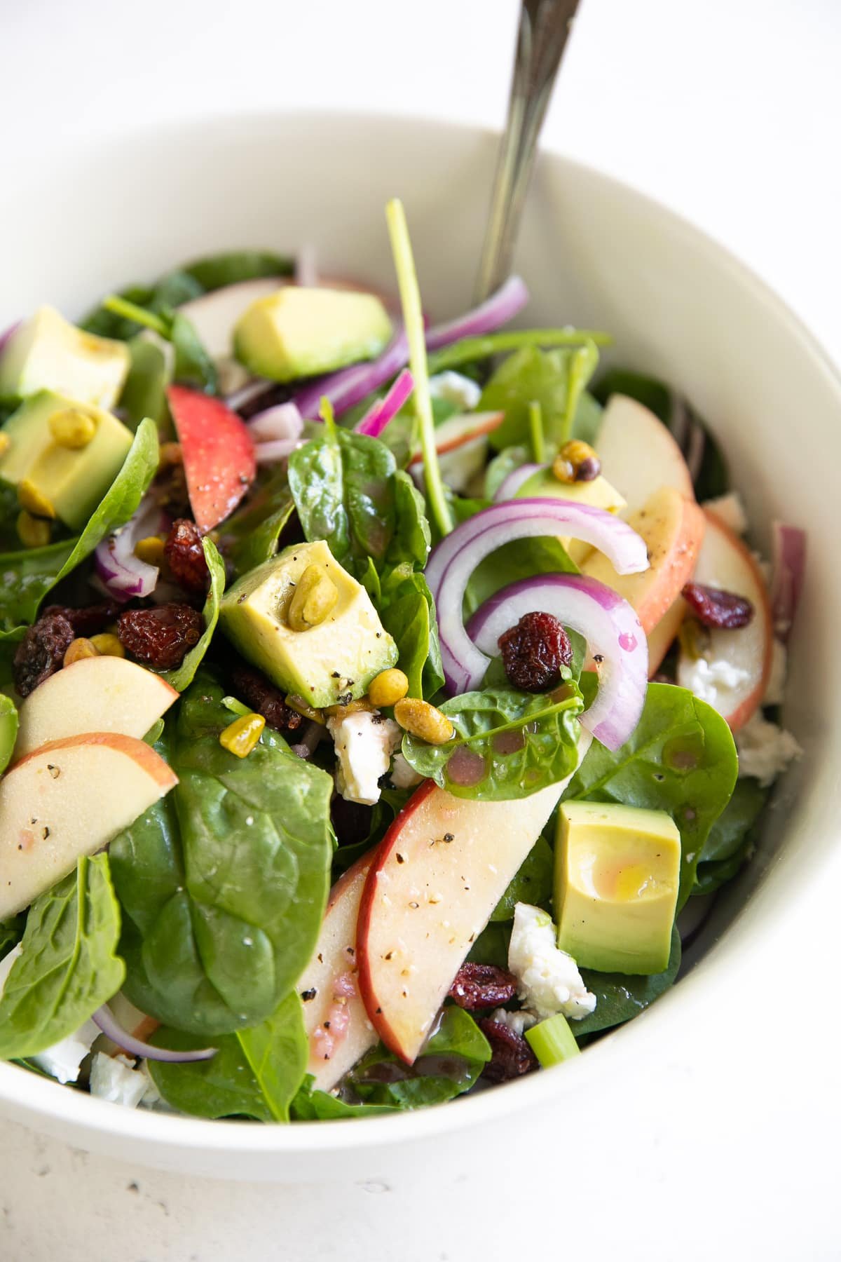 Large salad bowl filled with fresh baby spinach tossed with sliced apples, avocado, dried cranberries, feta cheese, and pistachios.