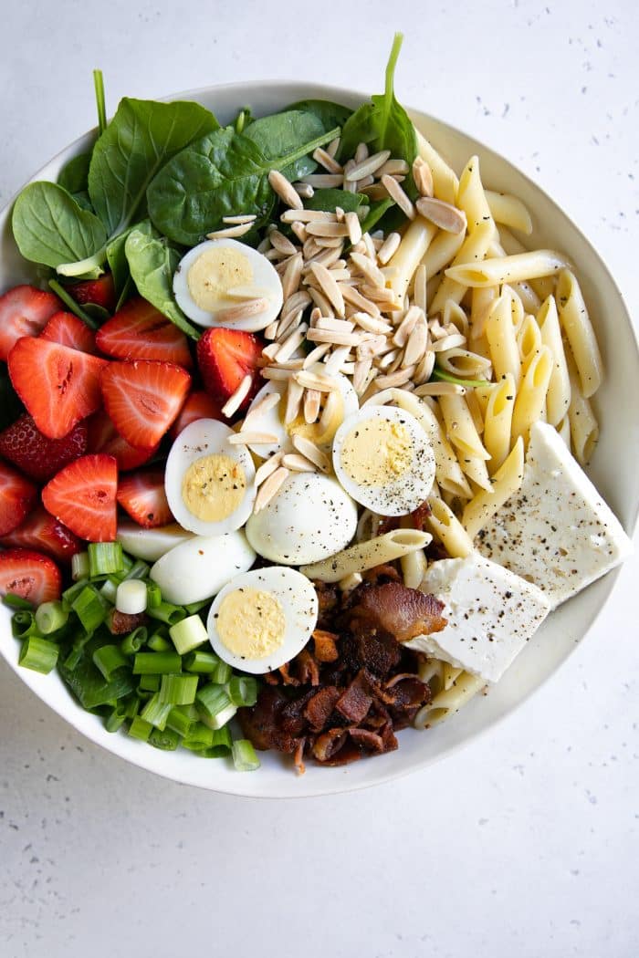 Large white salad bowl filled with penne, fresh spinach, feta cheese, strawberries, hard boiled eggs, and bacon.