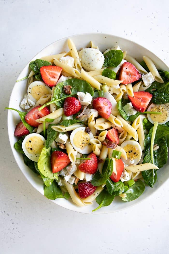 Large salad bowl filled with tossed together spinach pasta salad with strawberries, bacon, and hard-boiled eggs.