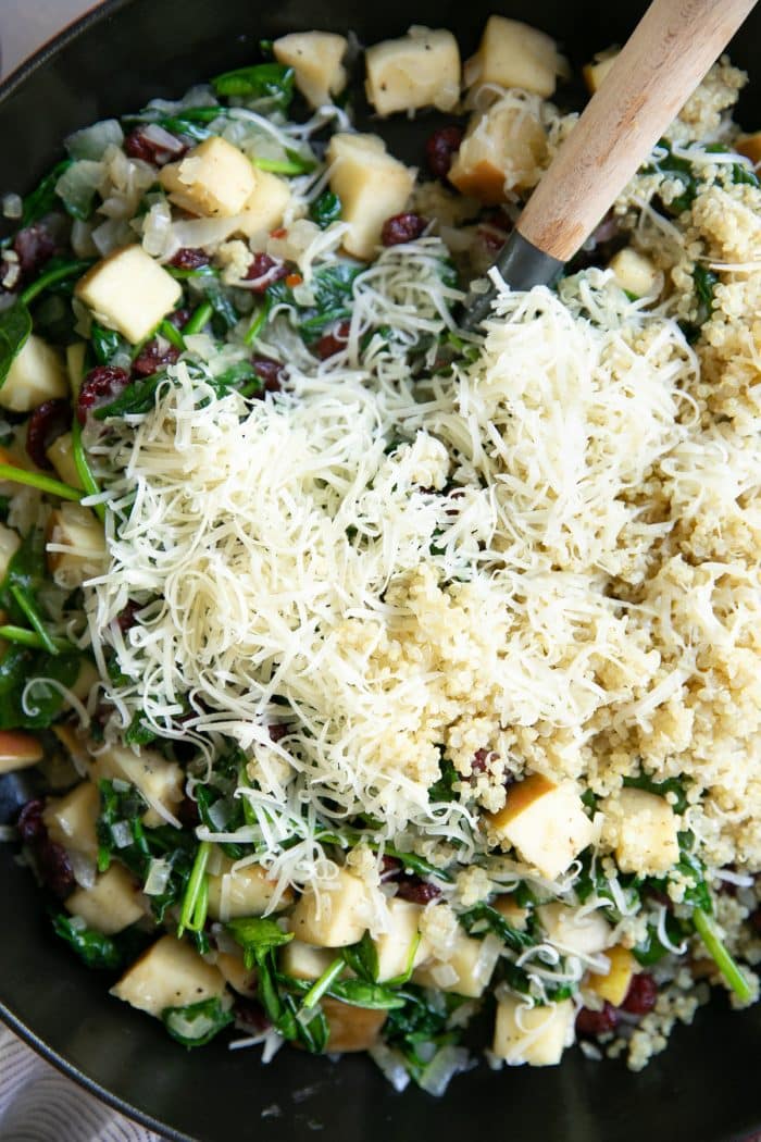 Skillet filled with cooked onions, apples, fresh sage, dried cranberries, spinach, and topped with cooked quinoa and shredded parmesan.