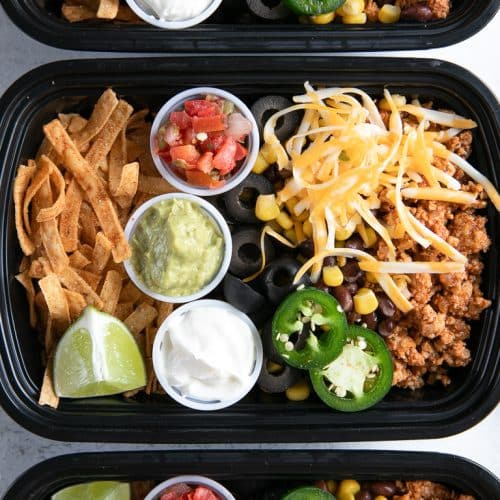 Three meal prep containers filled with seasoned ground turkey, cheese, olives, tortilla chips, and individual containers filled with sour cream, guacamole, and salsa.