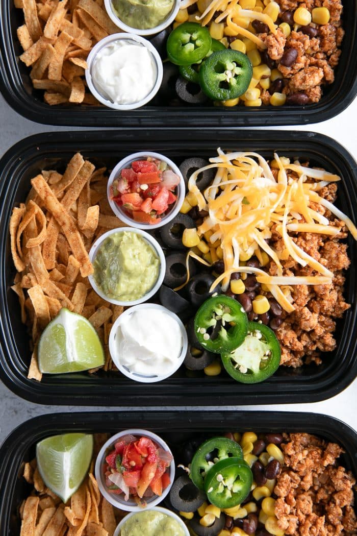 Three meal prep containers filled with seasoned ground turkey, cheese, olives, tortilla chips, and individual containers filled with sour cream, guacamole, and salsa.