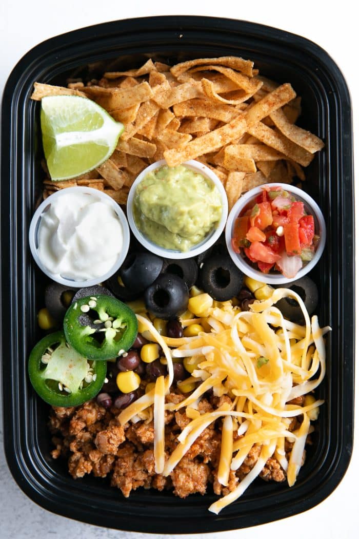 One meal prep container filled with seasoned ground turkey, cheese, olives, tortilla chips, and individual containers filled with sour cream, guacamole, and salsa.