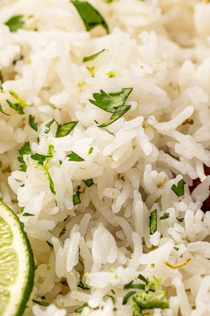 Close-up photo of fluffy white cilantro lime rice with visible chopped green herbs and thin strips of lime zest. Part of a lime wedge appears along the lower left edge, and the rice fills nearly the entire frame, highlighting the soft texture of the grains.