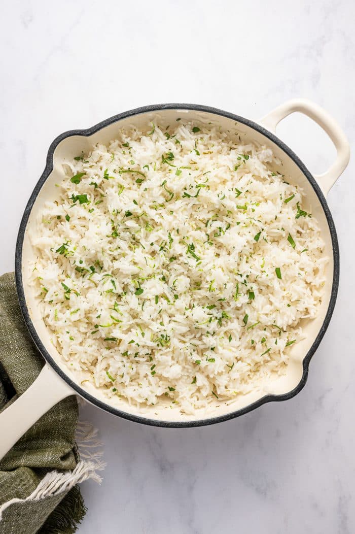 Overhead photo of freshly cooked cilantro lime rice in a round cream-colored pot with a black rim and side handles. The fluffy white rice is mixed with chopped green cilantro and lime zest and sits on a light gray marble background with an olive green fringed kitchen towel draped beneath the pot.