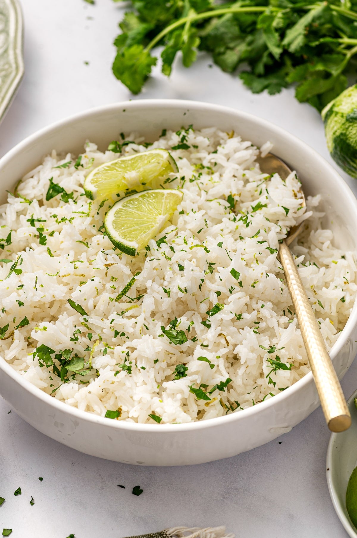 Angled close-up photo of cilantro lime rice in a round white bowl with a gold spoon resting along the right side. Two lime wedges sit on top of the rice, and chopped green herbs and fine lime zest are scattered throughout. Blurred fresh cilantro and lime appear in the background on a light gray marble surface.