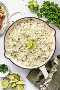 Overhead photo of fluffy cilantro lime rice in a round cream-colored pot with a black rim, garnished with two lime wedges in the center. Around the pot are fresh cilantro, a whole zested lime, a small white bowl of chopped herbs, a plate of lime wedges, part of a green patterned plate with sliced grilled chicken, and an olive green fringed kitchen towel on a light gray marble surface.