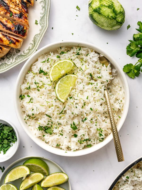 Overhead photo of cilantro lime rice served in a round white bowl with a gold spoon tucked into the right side and two lime wedges on top. The bowl is surrounded by a plate of lime wedges, a small white bowl of chopped herbs, fresh cilantro, a whole zested lime, part of a cream-colored pot, and a green patterned plate with sliced grilled chicken on a light gray marble surface.