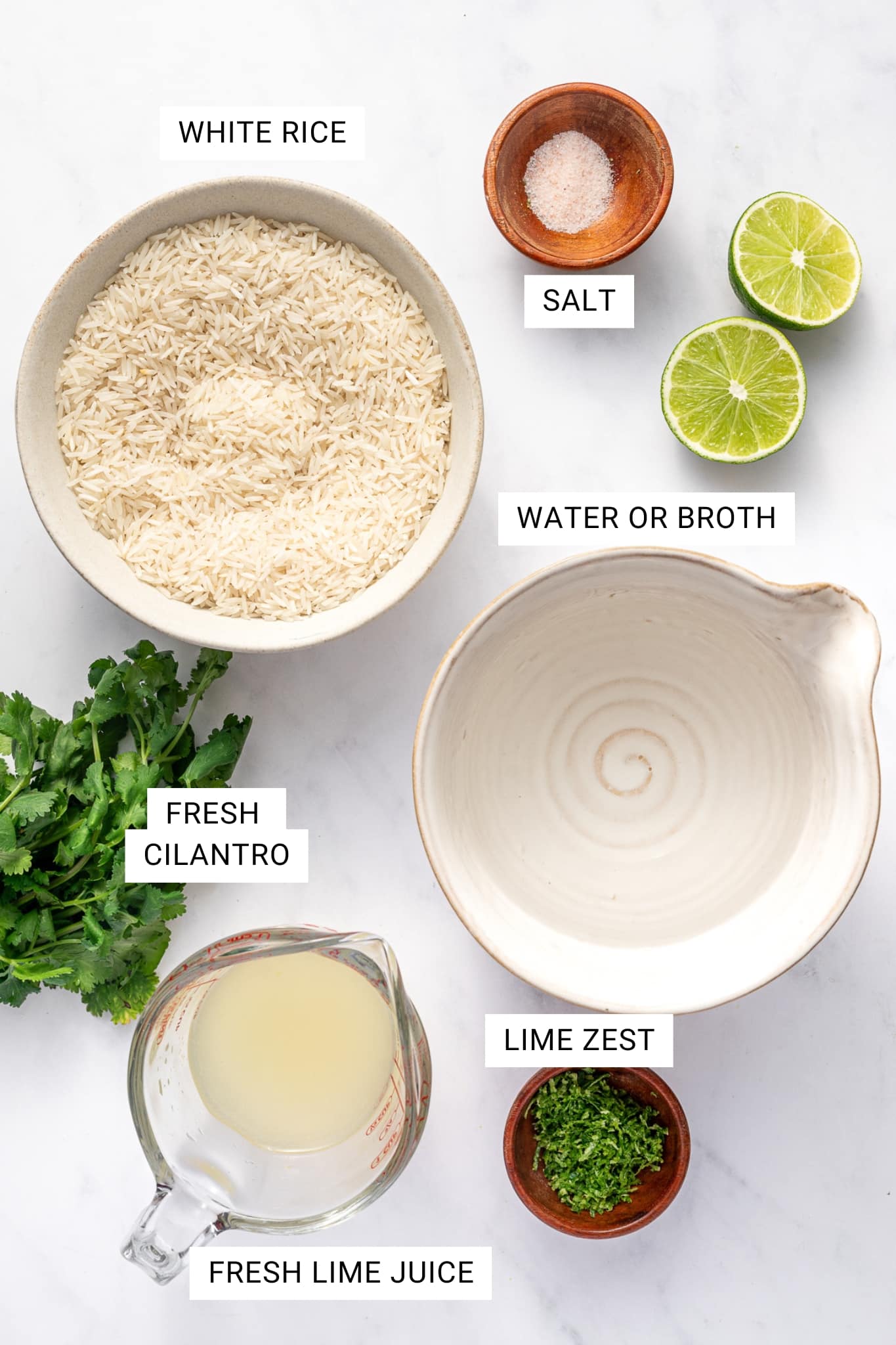 Overhead ingredient photo for cilantro lime rice on a light gray marble background with text overlays labeling each item. A beige bowl of uncooked white rice is labeled “WHITE RICE,” a small wooden bowl of salt is labeled “SALT,” an empty cream-colored bowl is labeled “WATER OR BROTH,” a bunch of fresh herbs at the lower left is labeled “FRESH CILANTRO,” a glass measuring cup of pale green juice is labeled “FRESH LIME JUICE,” and a small wooden bowl of green zest is labeled “LIME ZEST.” Two halved limes sit at the upper right.