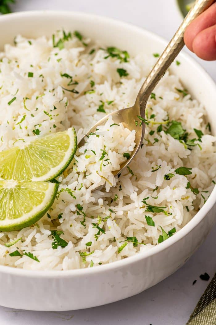 Close-up photo of a spoon lifting a bite of cilantro lime rice from a white bowl. The fluffy white rice is mixed with chopped green herbs and lime zest, and two lime wedges rest on top of the rice near the spoon. A hand is visible holding the spoon at the top right edge of the image.
