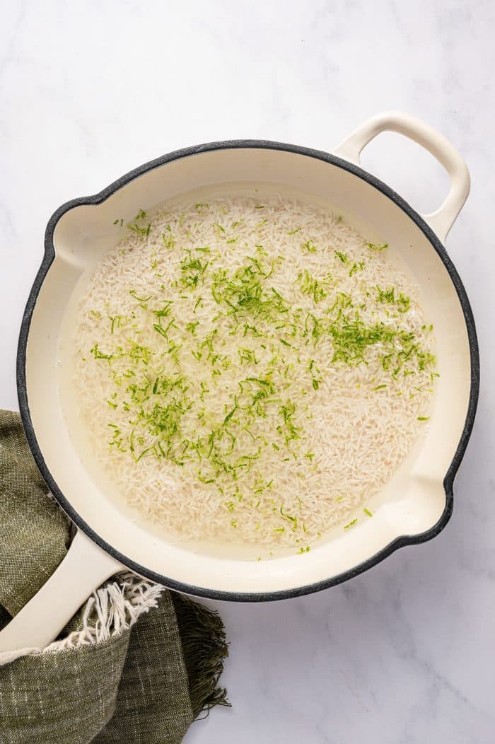 Overhead photo of uncooked white rice soaking in clear liquid inside a round cream-colored pot with a black rim and side handles, sprinkled with fresh green lime zest. The pot sits on a light gray marble surface with an olive green fringed kitchen towel tucked underneath the lower left side.