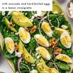 Pinterest pin for spinach salad on a light gray and food photography layout. A large white text box in the upper left corner contains black text that reads โSPINACH SALADโ with smaller black text below that reads โwith avocado and hard-boiled eggs in a lemon vinaigrette.โ The main image shows a large oval beige speckled platter filled with baby spinach, avocado slices, hard-boiled egg wedges, red onion, crumbled feta, walnuts, and black pepper. At the top right is a small white bowl of golden lemon vinaigrette, with a small beige bowl of feta nearby. A small bowl of walnuts appears at the bottom left, and an olive green napkin is styled along the right side. Theforkedspoon appears at the bottom.