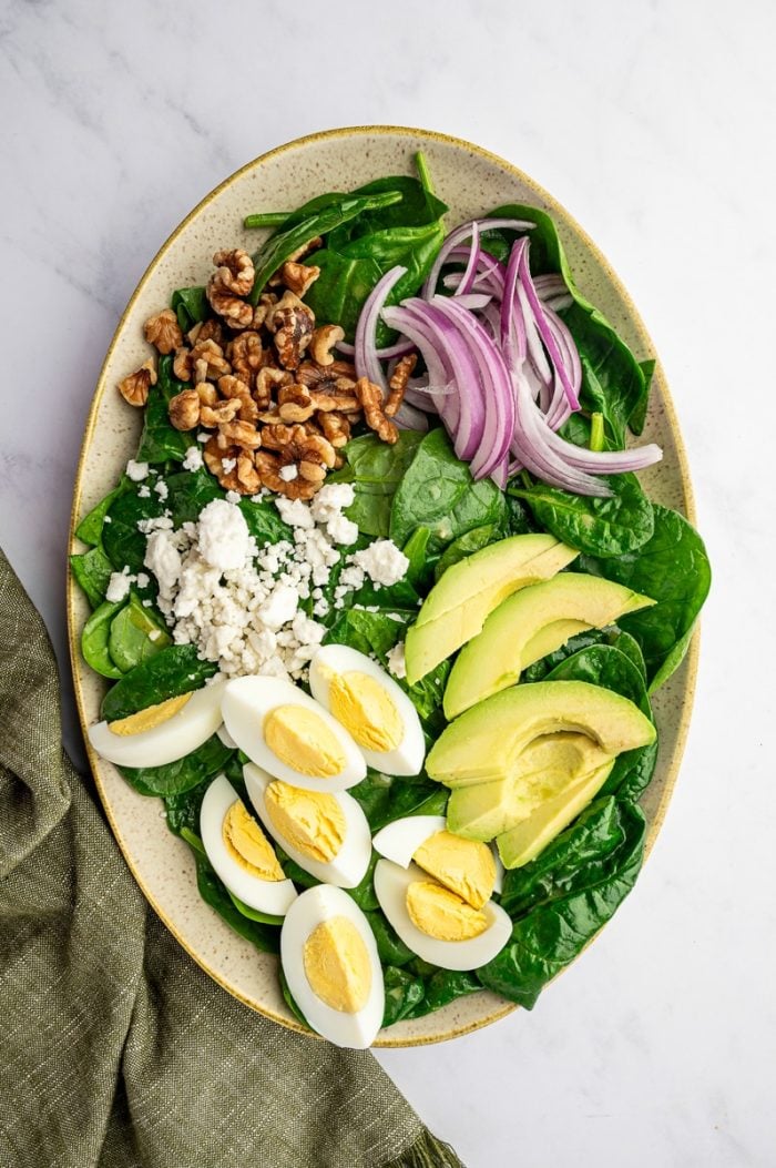 Spinach salad in progress in a large oval beige speckled platter with glossy baby spinach topped with sliced red onion, avocado wedges, hard-boiled egg quarters, crumbled feta cheese, and chopped walnuts on a white marble background with an olive green napkin at the lower right.