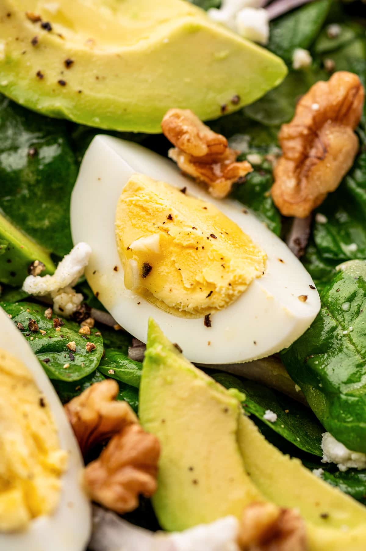 Close-up of spinach salad showing a hard-boiled egg wedge, avocado slices, baby spinach, walnut pieces, crumbled feta, and cracked black pepper; the image is tightly cropped to highlight texture and detail on the salad ingredients.