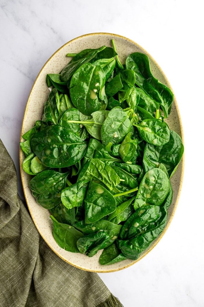 Baby spinach lightly tossed with dressing in a large oval beige speckled serving platter on a white marble surface with an olive green napkin at the lower right; the spinach leaves are glossy and coated with vinaigrette, with small bits of garlic visible on the leaves.