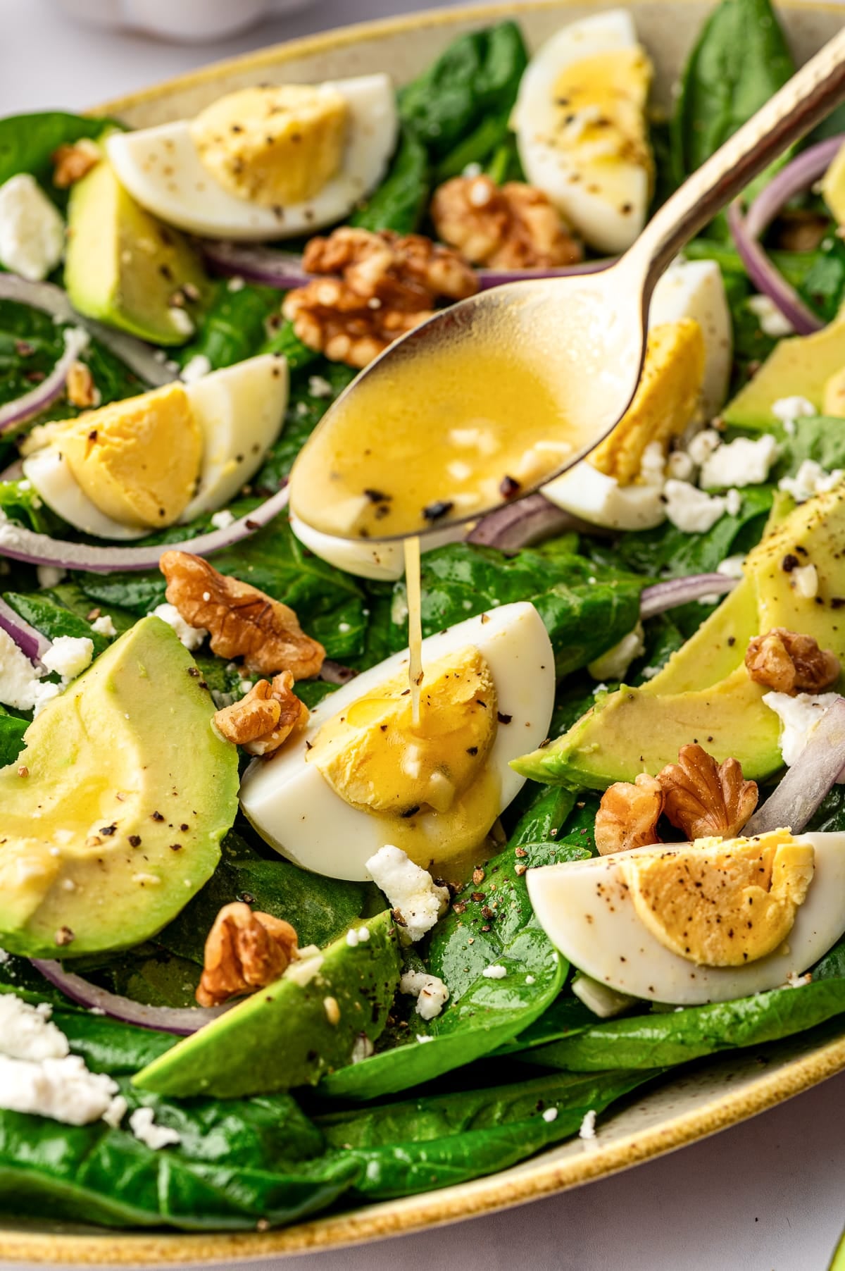 Gold spoon drizzling golden lemon vinaigrette with visible minced garlic and black pepper over a spinach salad topped with avocado slices, hard-boiled egg wedges, red onion, feta, and walnuts in a beige speckled platter; the salad sits on a white marble background.