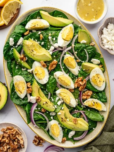 Overhead view of a finished spinach salad in a large oval beige speckled platter filled with baby spinach, avocado slices, hard-boiled egg wedges, thinly sliced red onion, crumbled feta, cracked black pepper, and walnuts; surrounding props include a white bowl of lemon vinaigrette, a small bowl of feta, lemon wedges on a wooden board, half an avocado, a small bowl of walnuts, and an olive green napkin on a white marble surface.