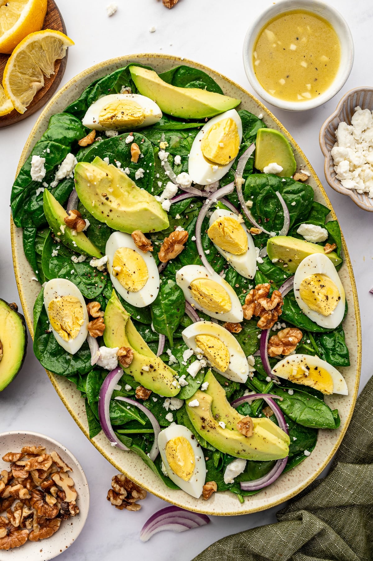 Overhead view of a finished spinach salad in a large oval beige speckled platter filled with baby spinach, avocado slices, hard-boiled egg wedges, thinly sliced red onion, crumbled feta, cracked black pepper, and walnuts; surrounding props include a white bowl of lemon vinaigrette, a small bowl of feta, lemon wedges on a wooden board, half an avocado, a small bowl of walnuts, and an olive green napkin on a white marble surface.