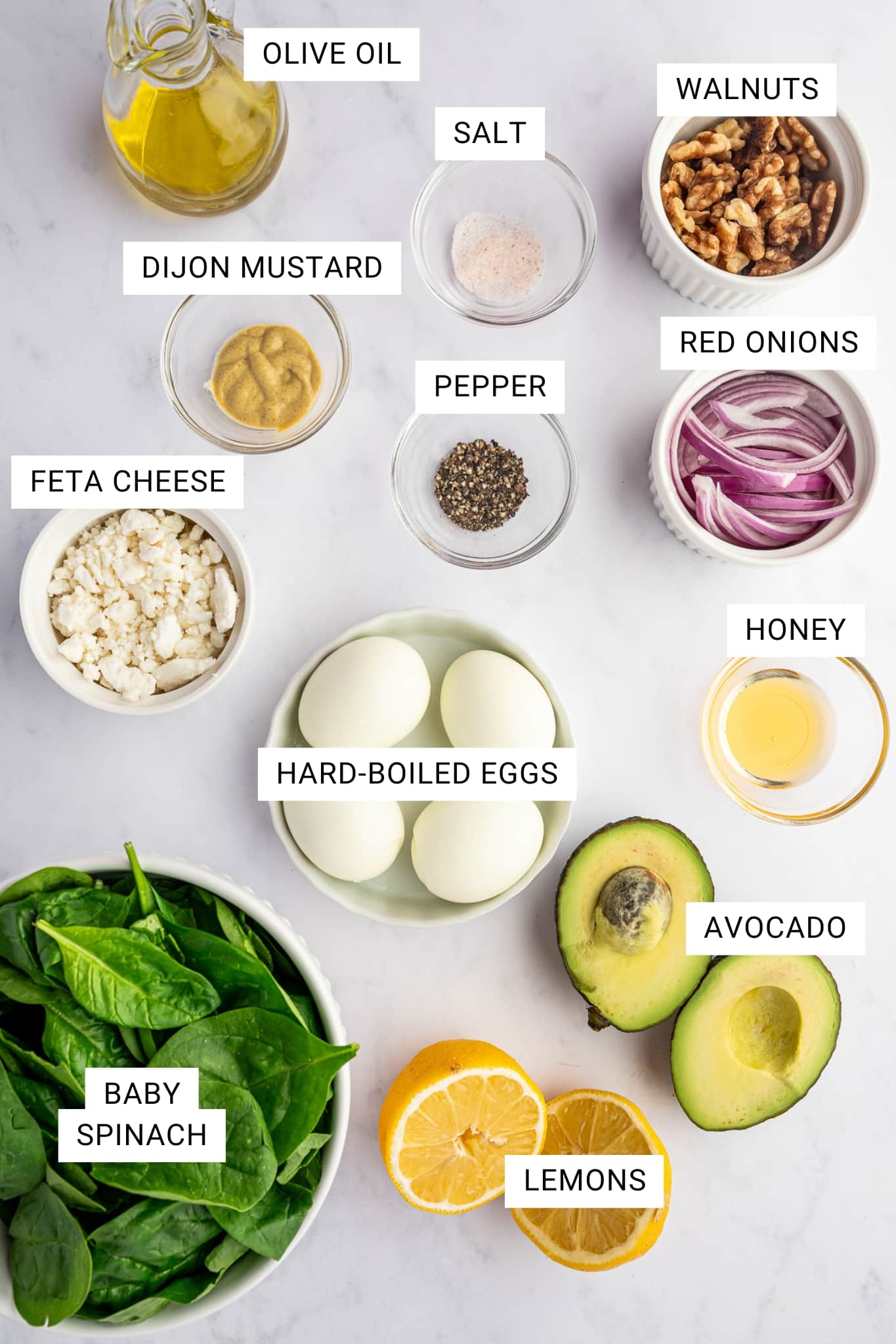 Ingredient shot for spinach salad on a white marble background with text overlays labeling each ingredient: OLIVE OIL, SALT, WALNUTS, DIJON MUSTARD, PEPPER, RED ONIONS, FETA CHEESE, HARD-BOILED EGGS, HONEY, BABY SPINACH, AVOCADO, and LEMONS; ingredients are arranged in bowls and ramekins with halved avocado and sliced lemons.