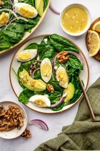 Individual serving of spinach salad in a shallow white bowl with a brown rim, topped with avocado slices, hard-boiled egg wedges, red onion, walnuts, feta, and black pepper; in the background are part of the larger serving platter, a small white bowl of lemon vinaigrette, lemon wedges on a wooden board, a small bowl of walnuts, and an olive green napkin on a white marble surface.