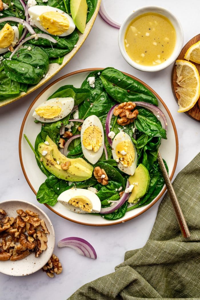 Individual serving of spinach salad in a shallow white bowl with a brown rim, topped with avocado slices, hard-boiled egg wedges, red onion, walnuts, feta, and black pepper; in the background are part of the larger serving platter, a small white bowl of lemon vinaigrette, lemon wedges on a wooden board, a small bowl of walnuts, and an olive green napkin on a white marble surface.