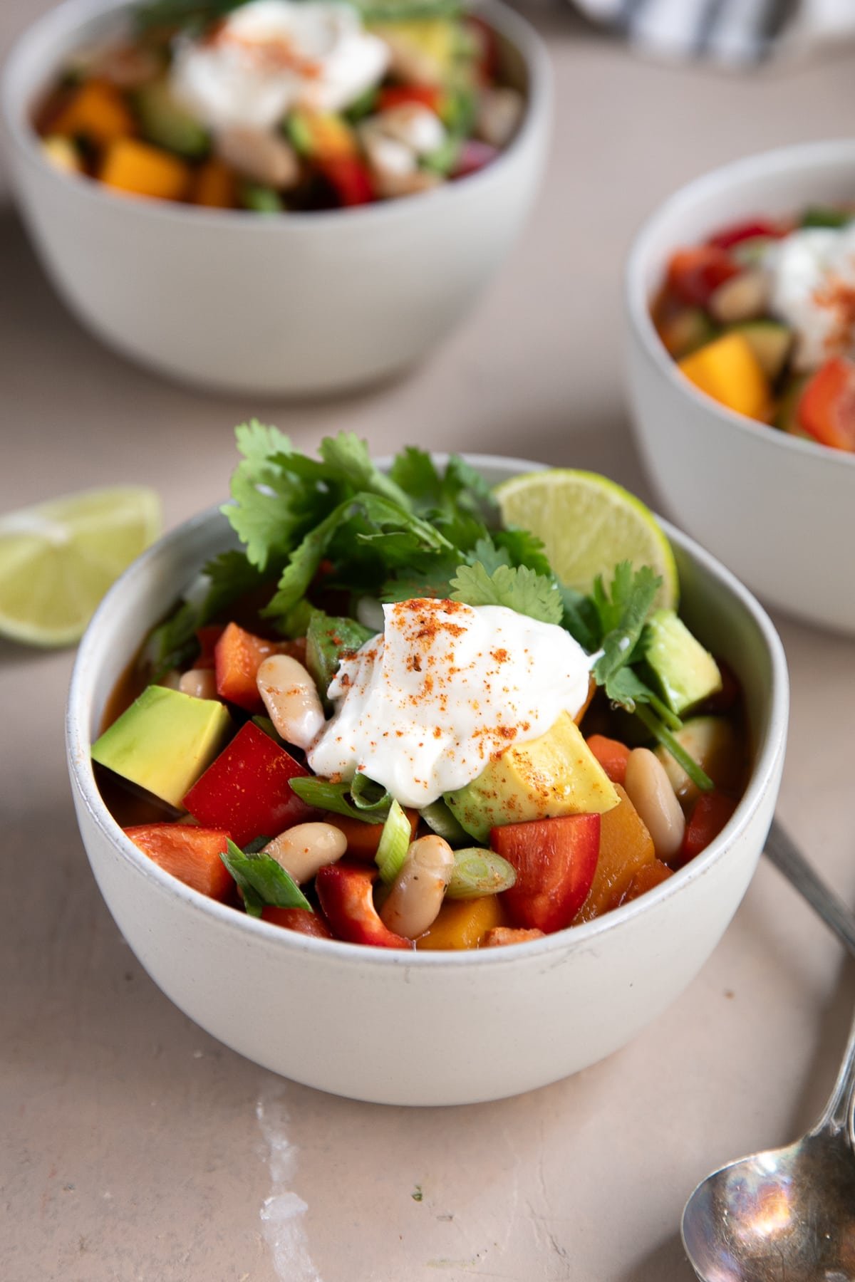 A close-up of a white bowl filled with colorful vegetarian butternut squash chili, topped with sour cream, sliced avocado, white beans, chopped green onions, fresh cilantro, and a lime wedge. The ingredients include red bell peppers, zucchini, and cubed butternut squash.