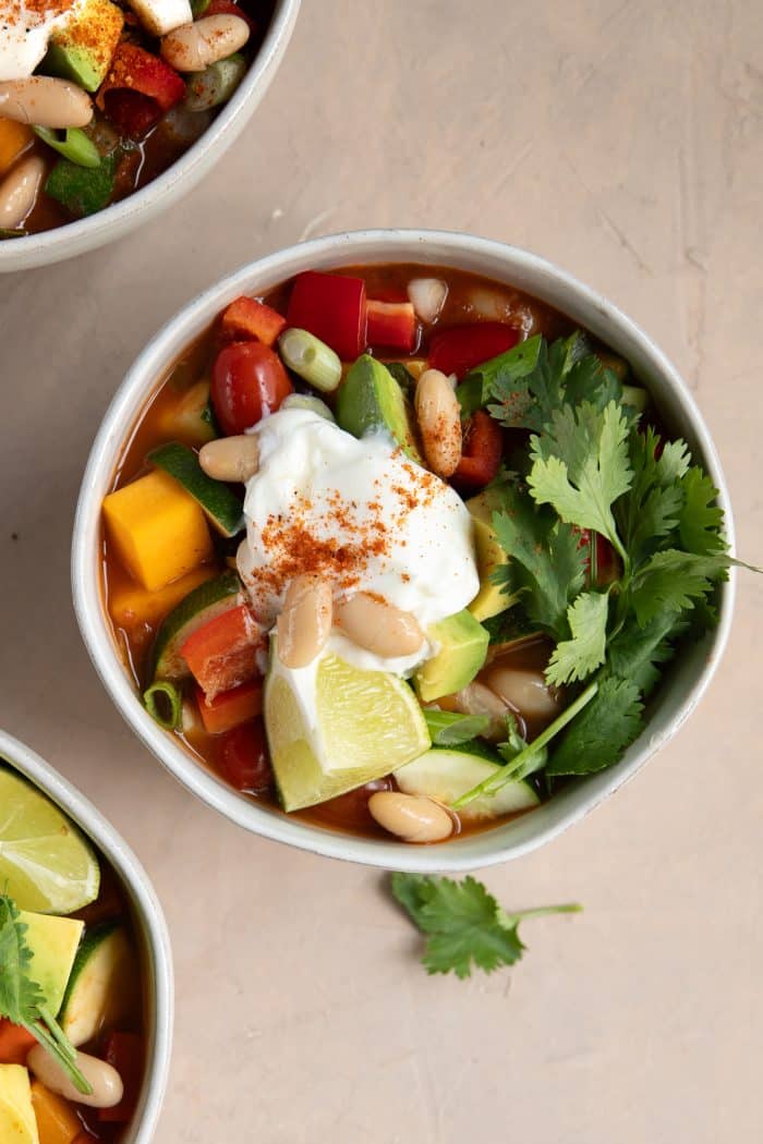 Overhead view of a bowl of vegetarian butternut squash chili, topped with a dollop of sour cream, cannellini beans, chopped avocado, lime wedges, and a generous handful of fresh cilantro.