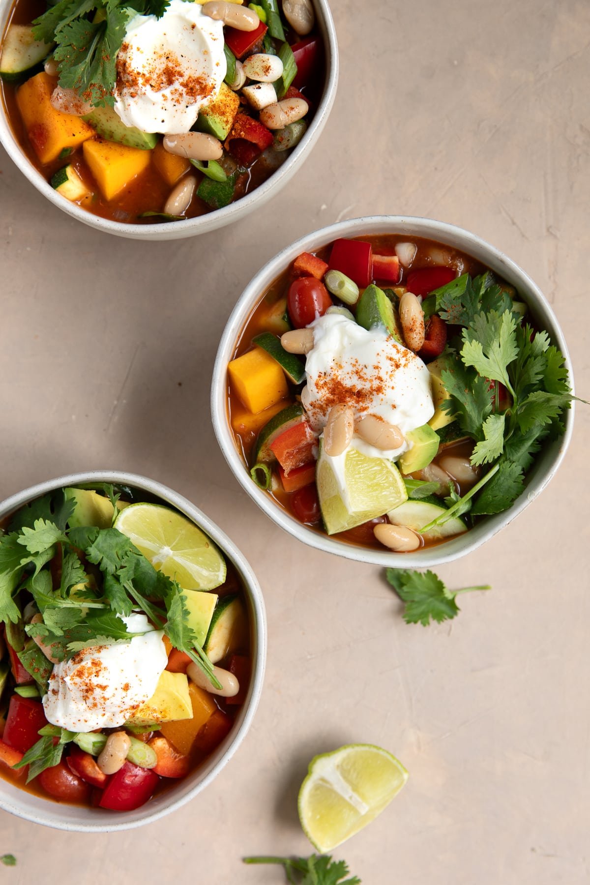 Three bowls of butternut squash chili arranged on a neutral surface, each garnished with sour cream, avocado, lime wedges, fresh cilantro, and a sprinkle of chili powder. A few loose cilantro leaves and lime wedges are scattered nearby.
