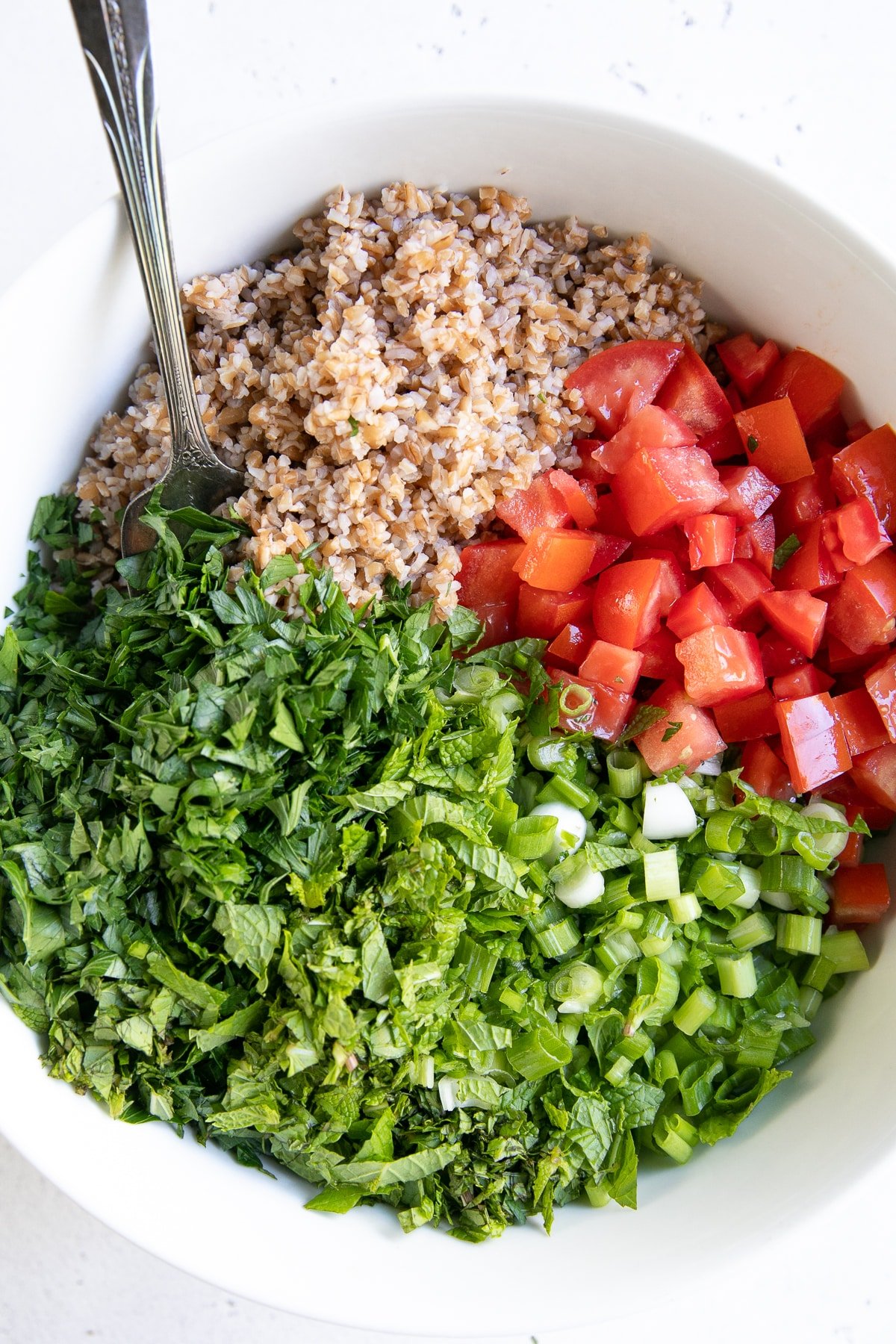 White salad bowl filled with cooked bulger, chopped tomatoes, green onion, mint, and parsley.