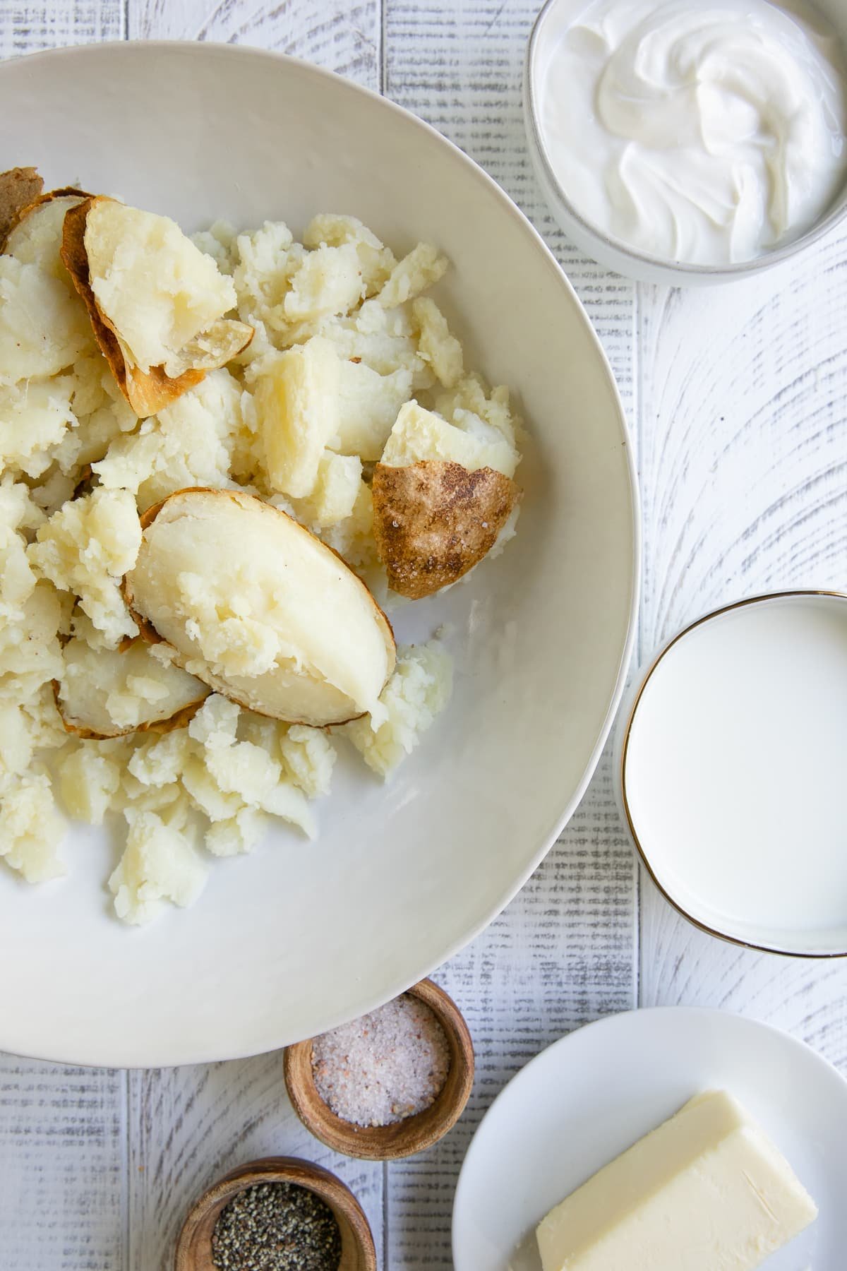 Large white bowl filled with the scooped out sides of baked potatoes and surrounded by side bowls of salt, pepper, sour cream, and milk.